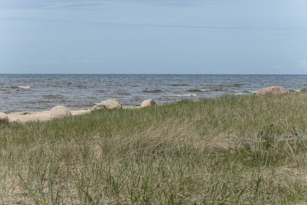 Темная скала, частично погруженная в морскую воду с маленькими волнами, разбивающимися на песчаном пляже под солнечным светом.