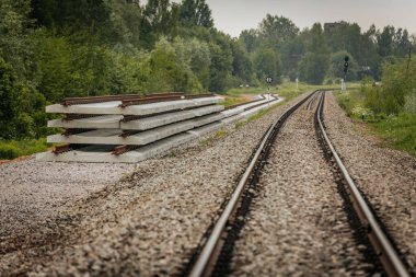 Düzgün istiflenmiş beton ve çelik demiryolu bölümleri yeşil ağaçlar ve bulutlu gökyüzü ile çevrili aktif bir demiryolu hattı boyunca konumlandırılmıştır..