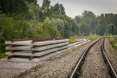 Düzgün istiflenmiş beton ve çelik demiryolu bölümleri yeşil ağaçlar ve bulutlu gökyüzü ile çevrili aktif bir demiryolu hattı boyunca konumlandırılmıştır..