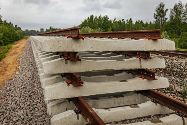 Düzgün istiflenmiş beton ve çelik demiryolu bölümleri yeşil ağaçlar ve bulutlu gökyüzü ile çevrili aktif bir demiryolu hattı boyunca konumlandırılmıştır..