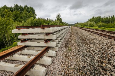 Düzgün istiflenmiş beton ve çelik demiryolu bölümleri yeşil ağaçlar ve bulutlu gökyüzü ile çevrili aktif bir demiryolu hattı boyunca konumlandırılmıştır..