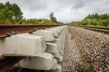 Düzgün istiflenmiş beton ve çelik demiryolu bölümleri yeşil ağaçlar ve bulutlu gökyüzü ile çevrili aktif bir demiryolu hattı boyunca konumlandırılmıştır..