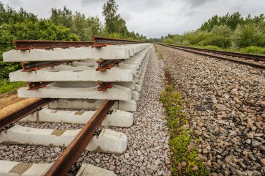 Düzgün istiflenmiş beton ve çelik demiryolu bölümleri yeşil ağaçlar ve bulutlu gökyüzü ile çevrili aktif bir demiryolu hattı boyunca konumlandırılmıştır..
