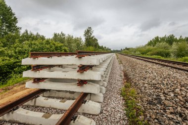 Düzgün istiflenmiş beton ve çelik demiryolu bölümleri yeşil ağaçlar ve bulutlu gökyüzü ile çevrili aktif bir demiryolu hattı boyunca konumlandırılmıştır..
