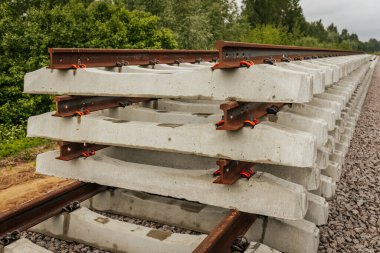 Düzgün istiflenmiş beton ve çelik demiryolu bölümleri yeşil ağaçlar ve bulutlu gökyüzü ile çevrili aktif bir demiryolu hattı boyunca konumlandırılmıştır..