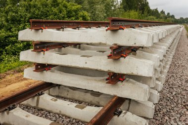 Düzgün istiflenmiş beton ve çelik demiryolu bölümleri yeşil ağaçlar ve bulutlu gökyüzü ile çevrili aktif bir demiryolu hattı boyunca konumlandırılmıştır..