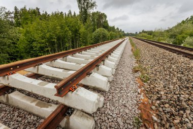 Düzgün istiflenmiş beton ve çelik demiryolu bölümleri yeşil ağaçlar ve bulutlu gökyüzü ile çevrili aktif bir demiryolu hattı boyunca konumlandırılmıştır..