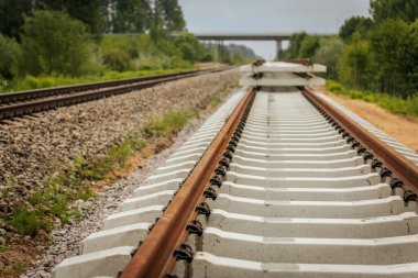 Düzgün istiflenmiş beton ve çelik demiryolu bölümleri yeşil ağaçlar ve bulutlu gökyüzü ile çevrili aktif bir demiryolu hattı boyunca konumlandırılmıştır..