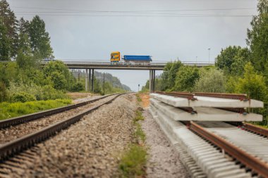 Düzgün istiflenmiş beton ve çelik demiryolu bölümleri yeşil ağaçlar ve bulutlu gökyüzü ile çevrili aktif bir demiryolu hattı boyunca konumlandırılmıştır..