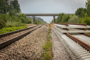 Düzgün istiflenmiş beton ve çelik demiryolu bölümleri yeşil ağaçlar ve bulutlu gökyüzü ile çevrili aktif bir demiryolu hattı boyunca konumlandırılmıştır..