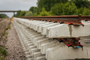 Düzgün istiflenmiş beton ve çelik demiryolu bölümleri yeşil ağaçlar ve bulutlu gökyüzü ile çevrili aktif bir demiryolu hattı boyunca konumlandırılmıştır..