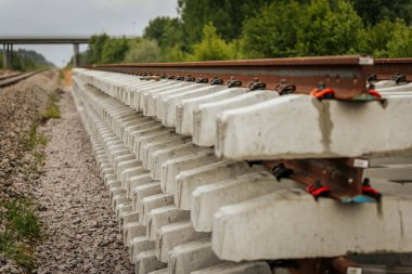 Düzgün istiflenmiş beton ve çelik demiryolu bölümleri yeşil ağaçlar ve bulutlu gökyüzü ile çevrili aktif bir demiryolu hattı boyunca konumlandırılmıştır..