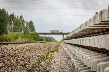 Düzgün istiflenmiş beton ve çelik demiryolu bölümleri yeşil ağaçlar ve bulutlu gökyüzü ile çevrili aktif bir demiryolu hattı boyunca konumlandırılmıştır..