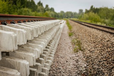 Düzgün istiflenmiş beton ve çelik demiryolu bölümleri yeşil ağaçlar ve bulutlu gökyüzü ile çevrili aktif bir demiryolu hattı boyunca konumlandırılmıştır..