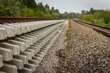 Düzgün istiflenmiş beton ve çelik demiryolu bölümleri yeşil ağaçlar ve bulutlu gökyüzü ile çevrili aktif bir demiryolu hattı boyunca konumlandırılmıştır..