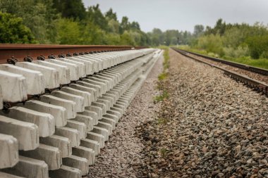Düzgün istiflenmiş beton ve çelik demiryolu bölümleri yeşil ağaçlar ve bulutlu gökyüzü ile çevrili aktif bir demiryolu hattı boyunca konumlandırılmıştır..