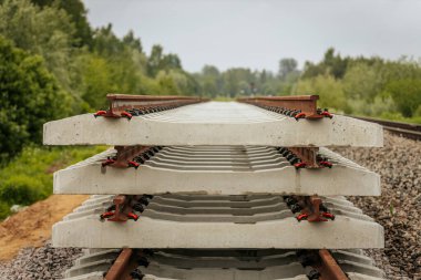 Düzgün istiflenmiş beton ve çelik demiryolu bölümleri yeşil ağaçlar ve bulutlu gökyüzü ile çevrili aktif bir demiryolu hattı boyunca konumlandırılmıştır..