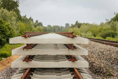 Düzgün istiflenmiş beton ve çelik demiryolu bölümleri yeşil ağaçlar ve bulutlu gökyüzü ile çevrili aktif bir demiryolu hattı boyunca konumlandırılmıştır..