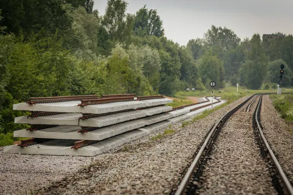 Düzgün istiflenmiş beton ve çelik demiryolu bölümleri yeşil ağaçlar ve bulutlu gökyüzü ile çevrili aktif bir demiryolu hattı boyunca konumlandırılmıştır..