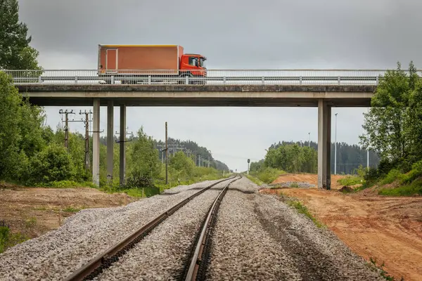 Uzun, düz tren yolları bulutlu bir gökyüzü altında yeşil orman alanlarının arasına kadar uzanıyor ve güçlü bir perspektif ve simetri sunuyor..