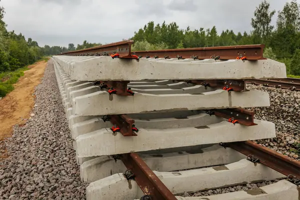 Düzgün istiflenmiş beton ve çelik demiryolu bölümleri yeşil ağaçlar ve bulutlu gökyüzü ile çevrili aktif bir demiryolu hattı boyunca konumlandırılmıştır..