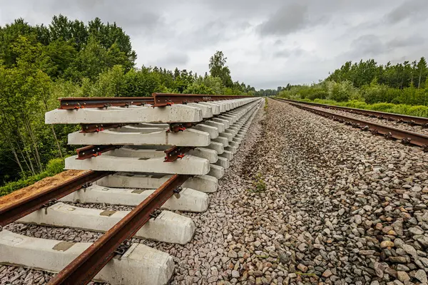 Düzgün istiflenmiş beton ve çelik demiryolu bölümleri yeşil ağaçlar ve bulutlu gökyüzü ile çevrili aktif bir demiryolu hattı boyunca konumlandırılmıştır..