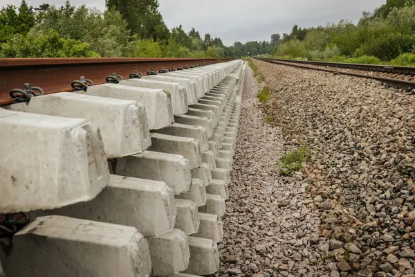 Düzgün istiflenmiş beton ve çelik demiryolu bölümleri yeşil ağaçlar ve bulutlu gökyüzü ile çevrili aktif bir demiryolu hattı boyunca konumlandırılmıştır..