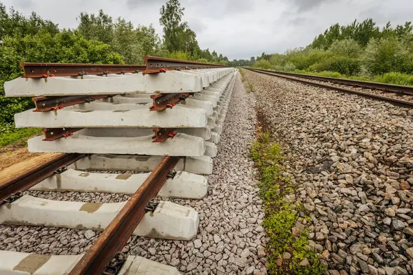 Düzgün istiflenmiş beton ve çelik demiryolu bölümleri yeşil ağaçlar ve bulutlu gökyüzü ile çevrili aktif bir demiryolu hattı boyunca konumlandırılmıştır..
