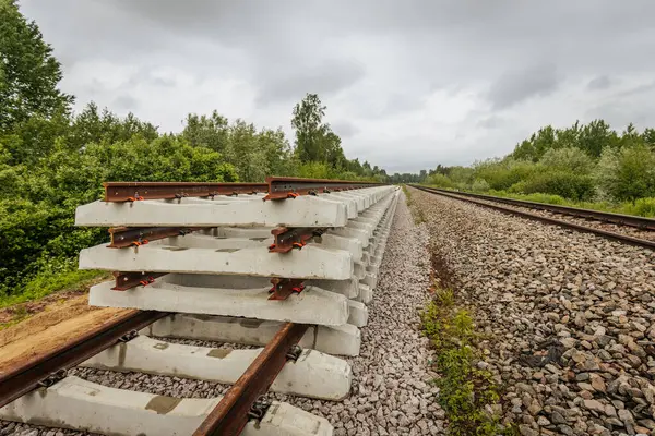Düzgün istiflenmiş beton ve çelik demiryolu bölümleri yeşil ağaçlar ve bulutlu gökyüzü ile çevrili aktif bir demiryolu hattı boyunca konumlandırılmıştır..