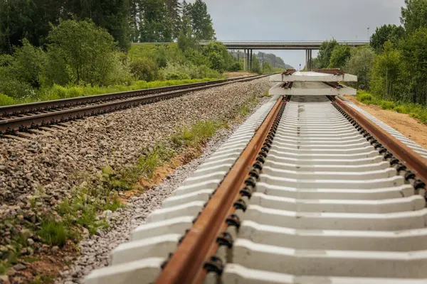 Düzgün istiflenmiş beton ve çelik demiryolu bölümleri yeşil ağaçlar ve bulutlu gökyüzü ile çevrili aktif bir demiryolu hattı boyunca konumlandırılmıştır..