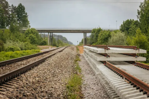 Düzgün istiflenmiş beton ve çelik demiryolu bölümleri yeşil ağaçlar ve bulutlu gökyüzü ile çevrili aktif bir demiryolu hattı boyunca konumlandırılmıştır..