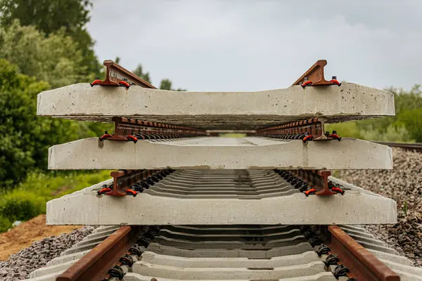 Düzgün istiflenmiş beton ve çelik demiryolu bölümleri yeşil ağaçlar ve bulutlu gökyüzü ile çevrili aktif bir demiryolu hattı boyunca konumlandırılmıştır..
