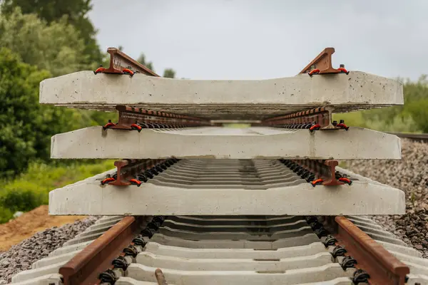 Düzgün istiflenmiş beton ve çelik demiryolu bölümleri yeşil ağaçlar ve bulutlu gökyüzü ile çevrili aktif bir demiryolu hattı boyunca konumlandırılmıştır..