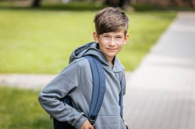 Mavi bir sırt çantası olan gri kapüşonlu çocuk kaldırımlı bir yolda duruyor, elleri ceplerinde, hafifçe gülümseyerek yan tarafa bakıyor..