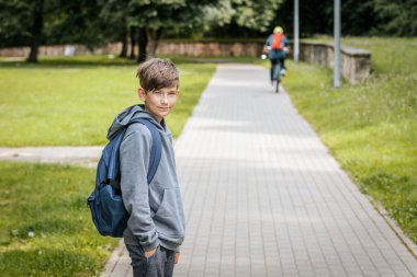 Mavi bir sırt çantası olan gri kapüşonlu çocuk kaldırımlı bir yolda duruyor, elleri ceplerinde, hafifçe gülümseyerek yan tarafa bakıyor..