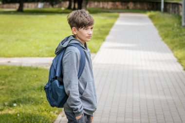 Mavi bir sırt çantası olan gri kapüşonlu çocuk kaldırımlı bir yolda duruyor, elleri ceplerinde, hafifçe gülümseyerek yan tarafa bakıyor..