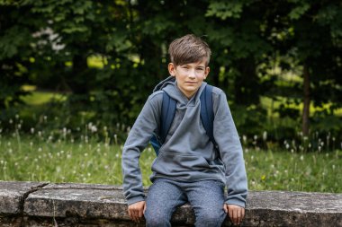 Gri kapüşonlu, mavi çantalı bir çocuk çimenli bir alanda duruyor, bir eli kalçasında duruyor ve kameraya bakıyor..