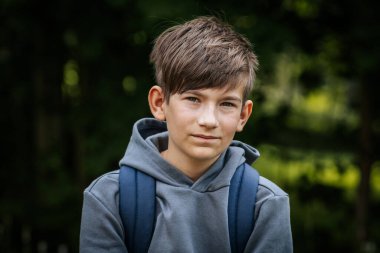 Gri kapüşonlu, mavi çantalı bir çocuk çimenli bir alanda duruyor, bir eli kalçasında duruyor ve kameraya bakıyor..