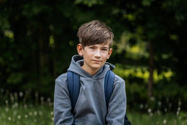 Gri kapüşonlu, mavi çantalı bir çocuk çimenli bir alanda duruyor, bir eli kalçasında duruyor ve kameraya bakıyor..