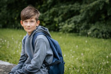 Gri kapüşonlu, mavi çantalı bir çocuk çimenli bir alanda duruyor, bir eli kalçasında duruyor ve kameraya bakıyor..