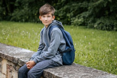 Gri kapüşonlu, mavi çantalı bir çocuk çimenli bir alanda duruyor, bir eli kalçasında duruyor ve kameraya bakıyor..