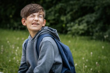 Gri kapüşonlu, mavi çantalı bir çocuk çimenli bir alanda duruyor, bir eli kalçasında duruyor ve kameraya bakıyor..