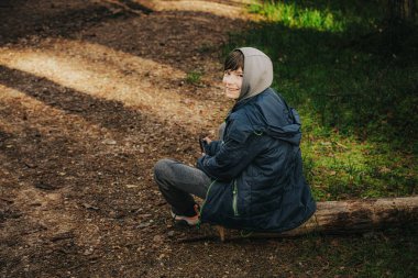 Smiling boy in a hoodie and jacket sits on a fallen log along a forest path, enjoying nature and sunlight in the woods.