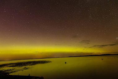 Bright green and red northern lights dance over a calm lake, casting vivid reflections on the water under a star-filled sky during a clear night.