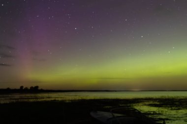 Bright green and red northern lights dance over a calm lake, casting vivid reflections on the water under a star-filled sky during a clear night.
