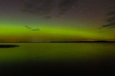 Bright green and red northern lights dance over a calm lake, casting vivid reflections on the water under a star-filled sky during a clear night.