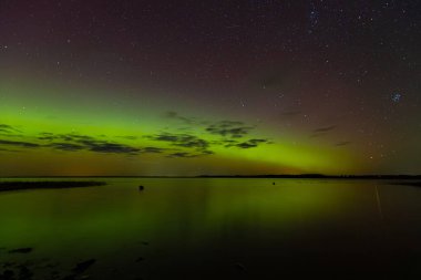 Bright green and red northern lights dance over a calm lake, casting vivid reflections on the water under a star-filled sky during a clear night.