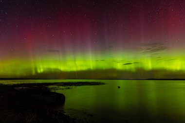 Bright green and red northern lights dance over a calm lake, casting vivid reflections on the water under a star-filled sky during a clear night.