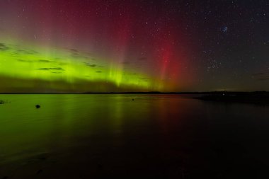 Bright green and red northern lights dance over a calm lake, casting vivid reflections on the water under a star-filled sky during a clear night.