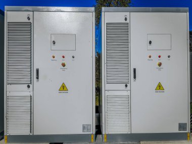 Açık hava yüksek voltajlı elektrikli dolaplar. Uyarı etiketleri, havalandırma panelleri ve açık mavi gökyüzünün altındaki acil durdurma düğmeleri..